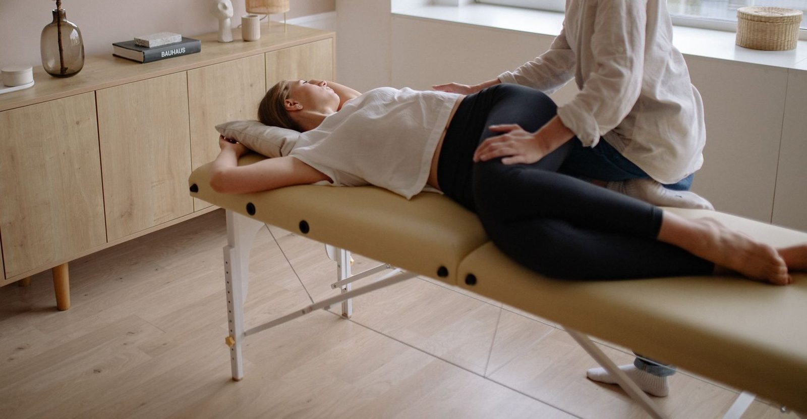 Fisioterapeuta tratando la cadera de una paciente tumbada, en una sala con luz natural.